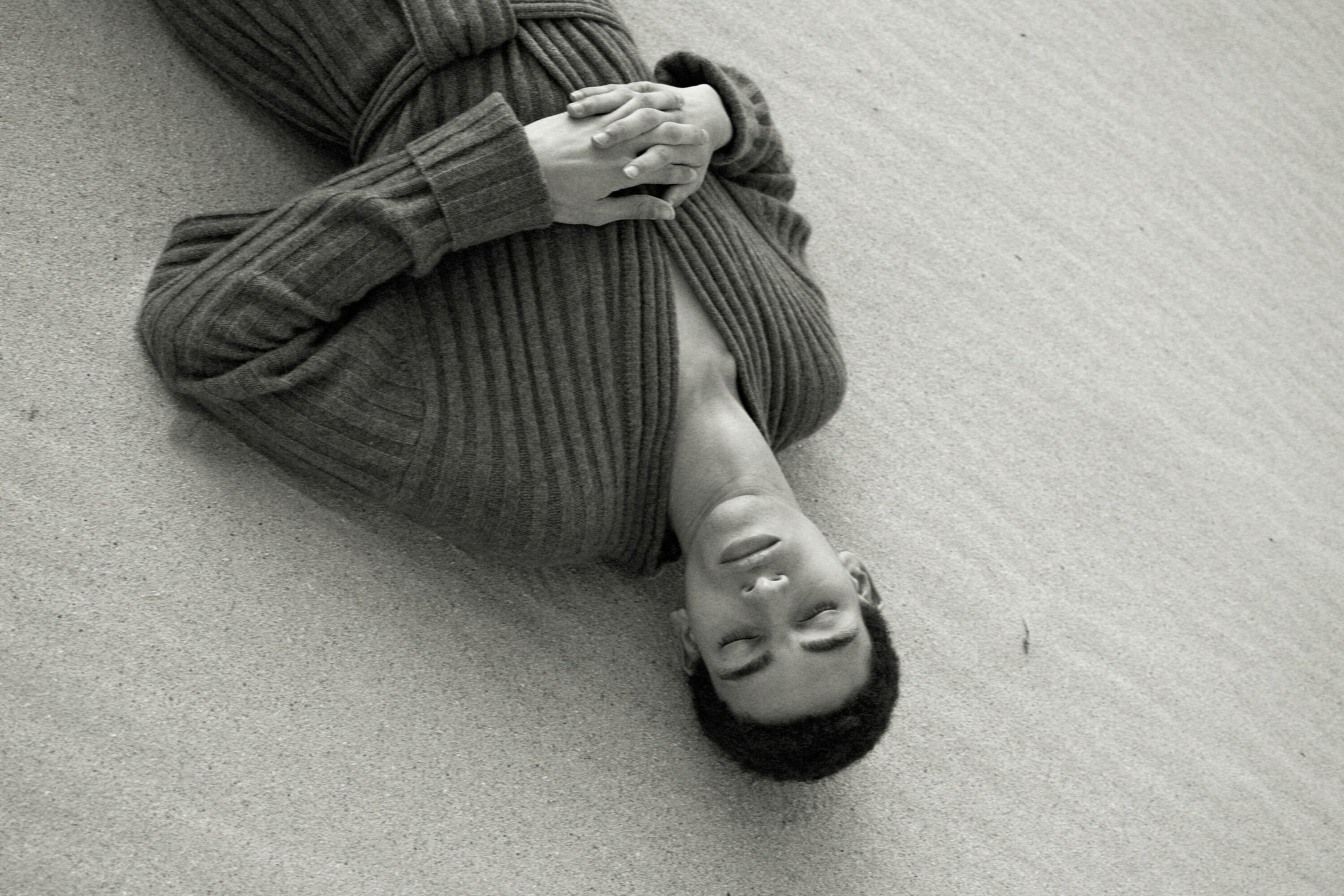 Young adult lies on textured surface wearing a gray robe in a monochrome shot. Hands are clasped, eyes are closed in a serene setting.