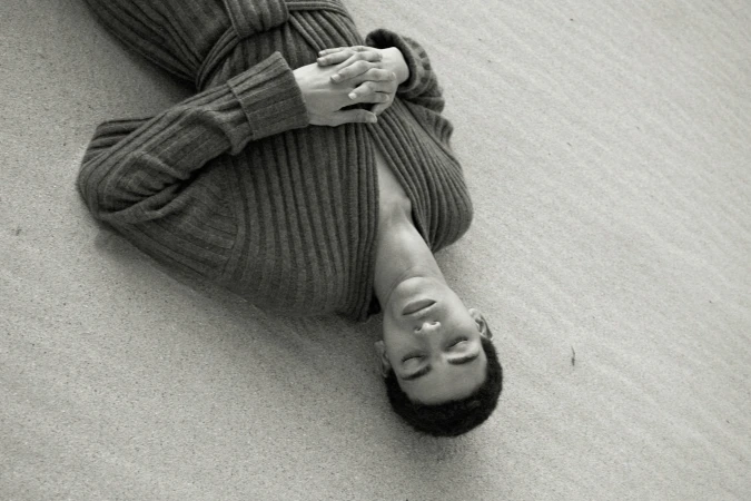 Young adult lies on textured surface wearing a gray robe in a monochrome shot. Hands are clasped, eyes are closed in a serene setting.