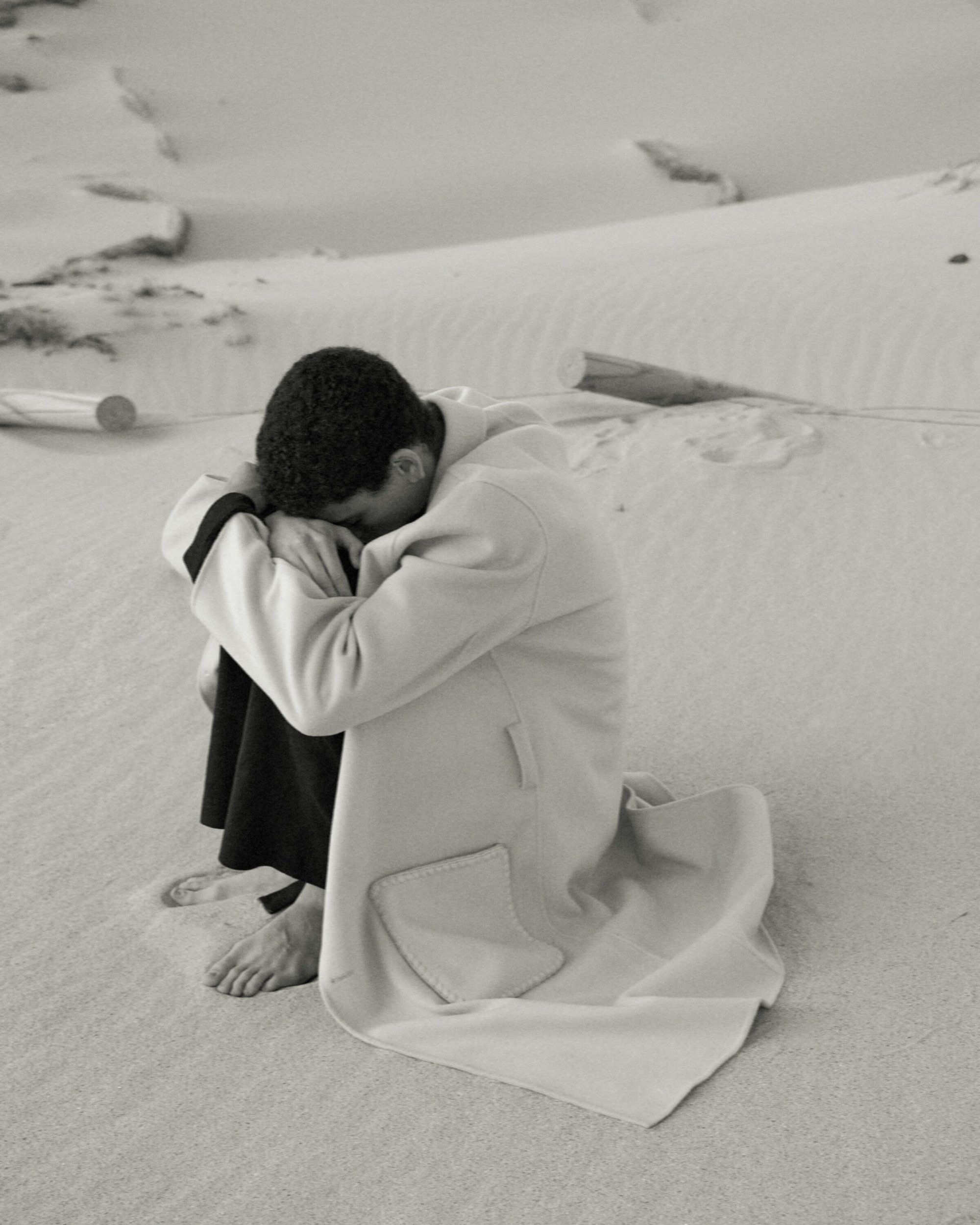 Monochrome portrait of a crouched person in a coat on a sandy beach, facing away with head down, near driftwood.