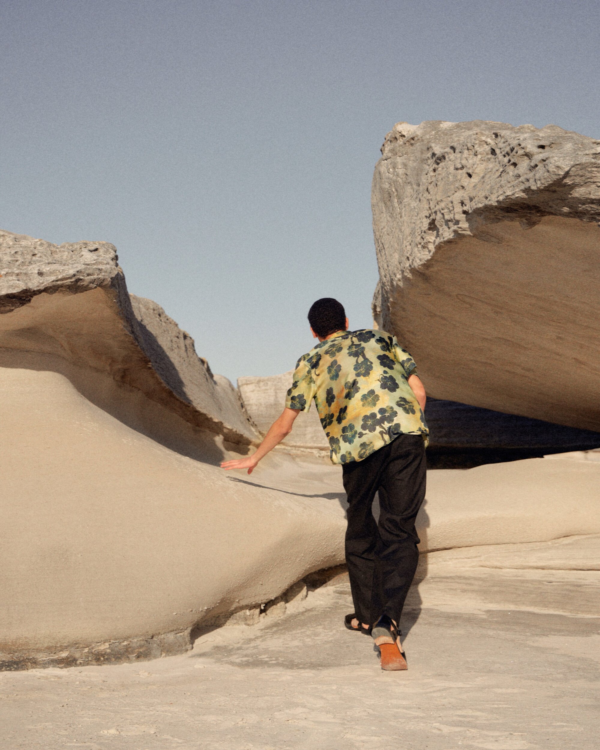A person wearing a floral shirt and dark pants walks across a light sandy ground toward large rock formations under a clear sky.