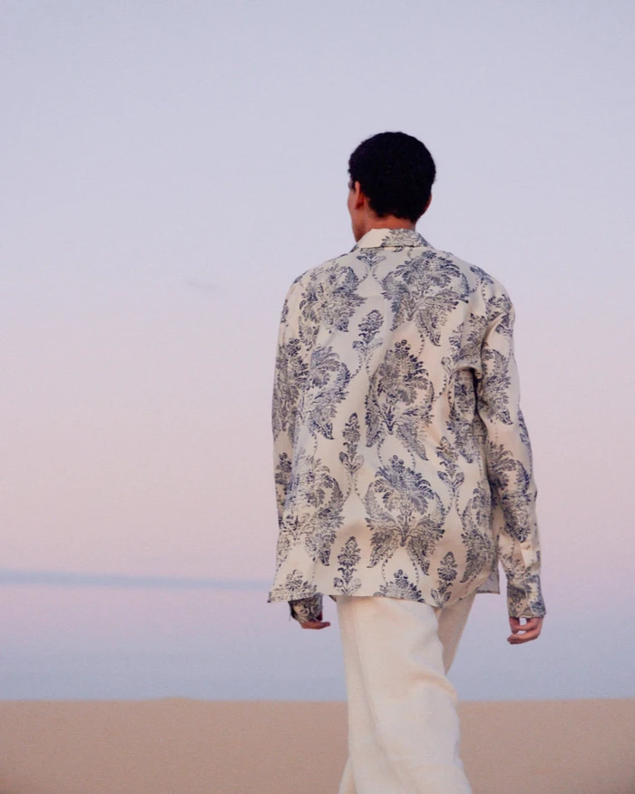 Man in a printed cream and blue long-sleeve shirt walking away across sand dunes under a pastel twilight sky.