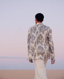 Man in a printed cream and blue long-sleeve shirt walking away across sand dunes under a pastel twilight sky.