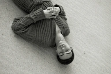 Model lying on sandy beach wearing a ribbed knit garment with hands folded across chest in black and white