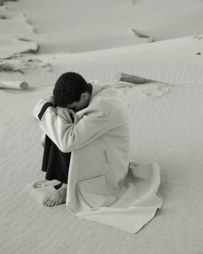 Model Sereto Boardman sitting curled up barefoot on a sandy dune wearing a long cream coat with black lining.