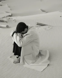 Model Sereto Boardman sitting curled up barefoot on a sandy dune wearing a long cream coat with black lining.