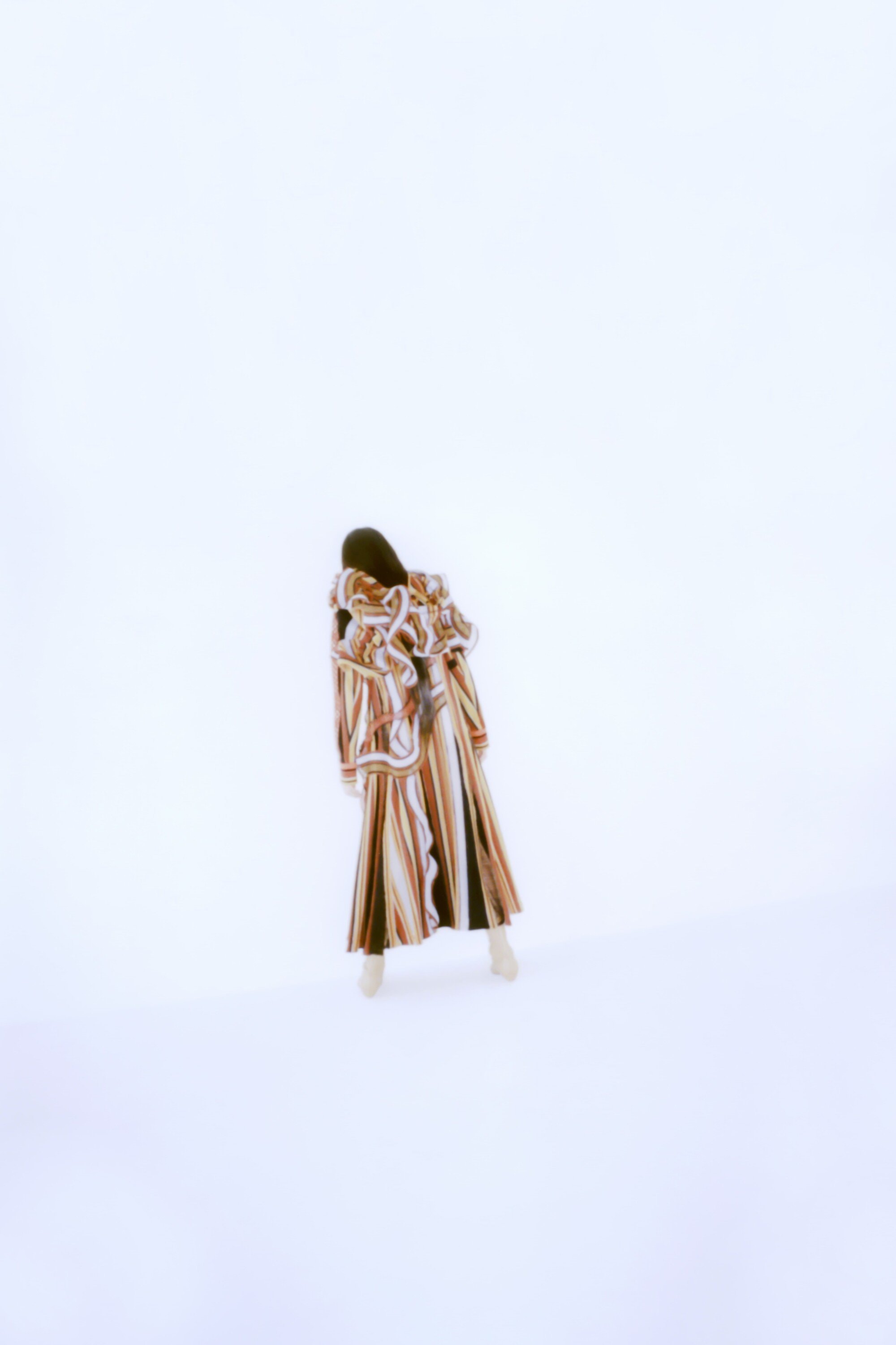 A single woman in a long patterned coat standing in a brightly lit white room, presenting a fashion-forward and artistic aesthetic.