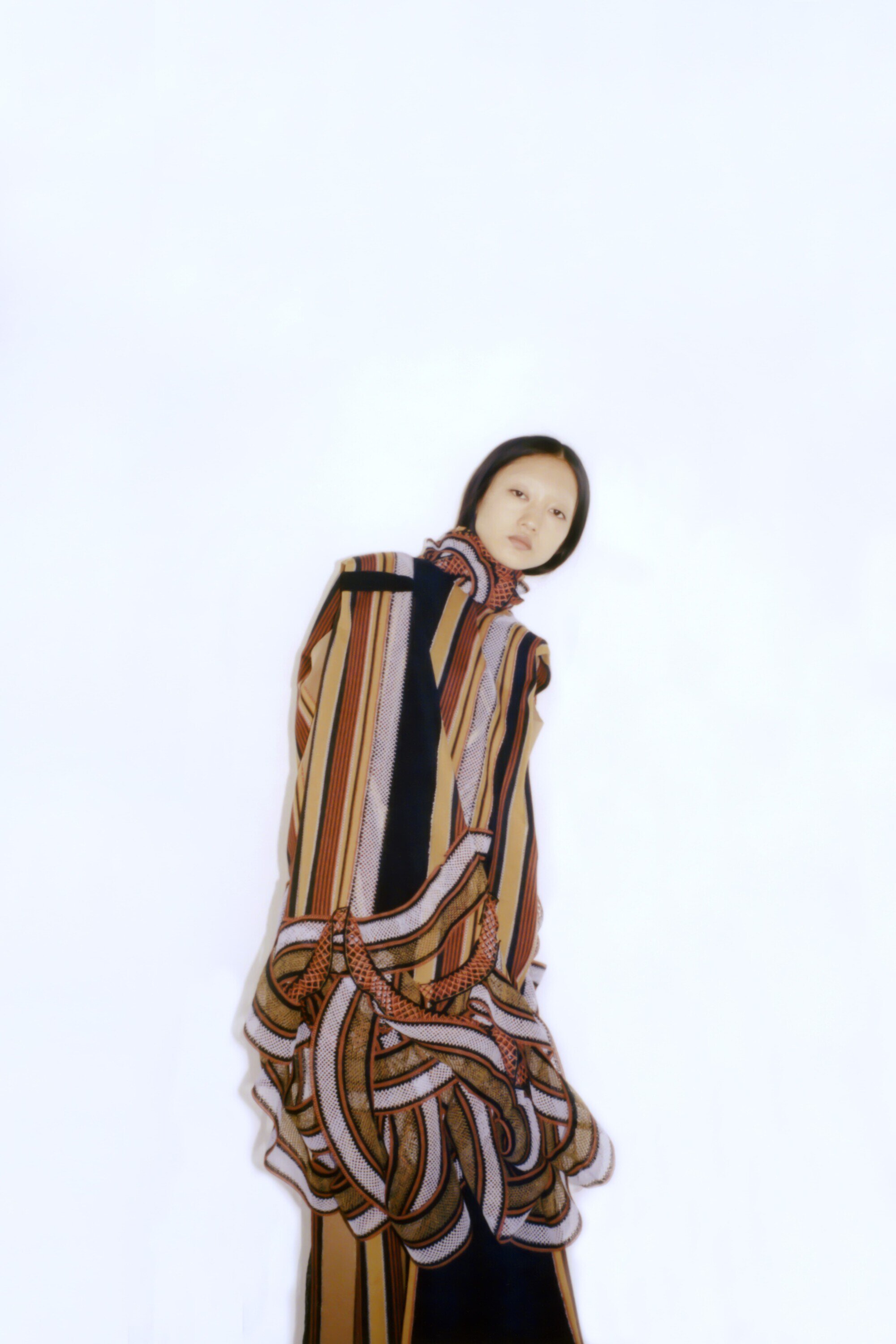 Asian woman in a striped coat posing against a white background. Fashion-forward design featuring earth tones, black, and a unique ruffle detail.