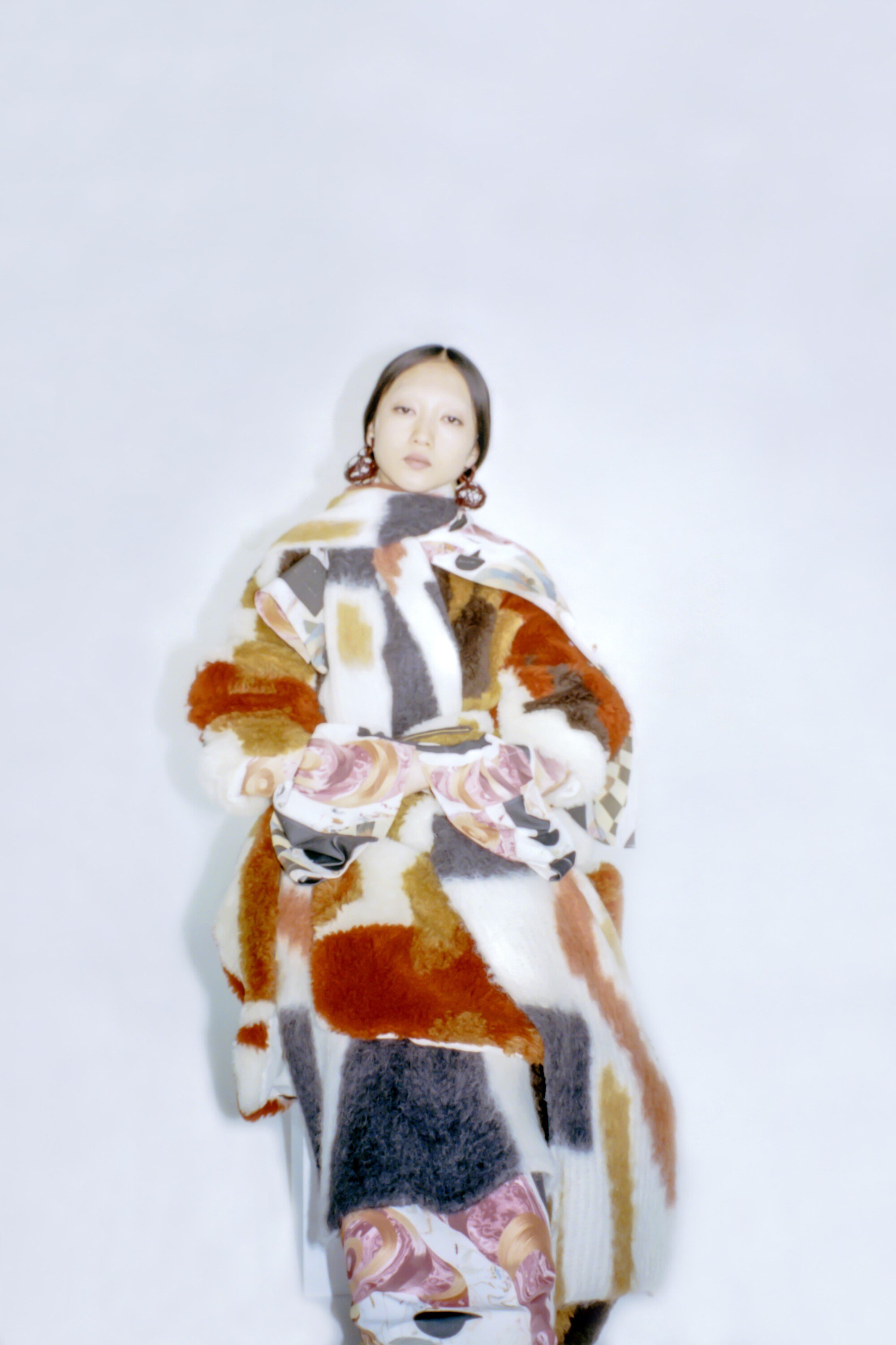 Woman in large multicolor coat with matching pants against a white background, wearing large round earrings. Fashion photoshoot.