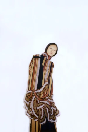 Model Mona Kawasaki wearing an earth-toned striped sculptural draped dress against a white background.