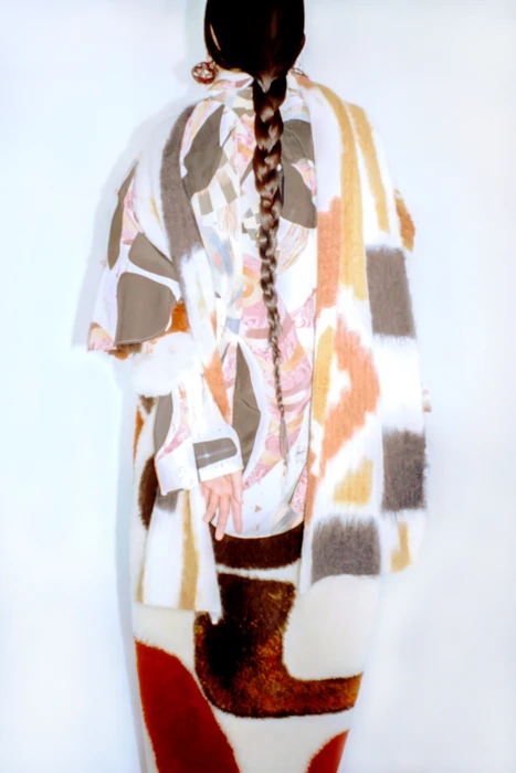 Model Mona Kawasaki viewed from behind with a long braided ponytail, wearing a textured abstract patterned coat against a white backdrop.