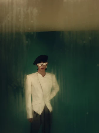 Model wearing a white structured blazer, black beret, and embellished sunglasses against a dark green textured background.