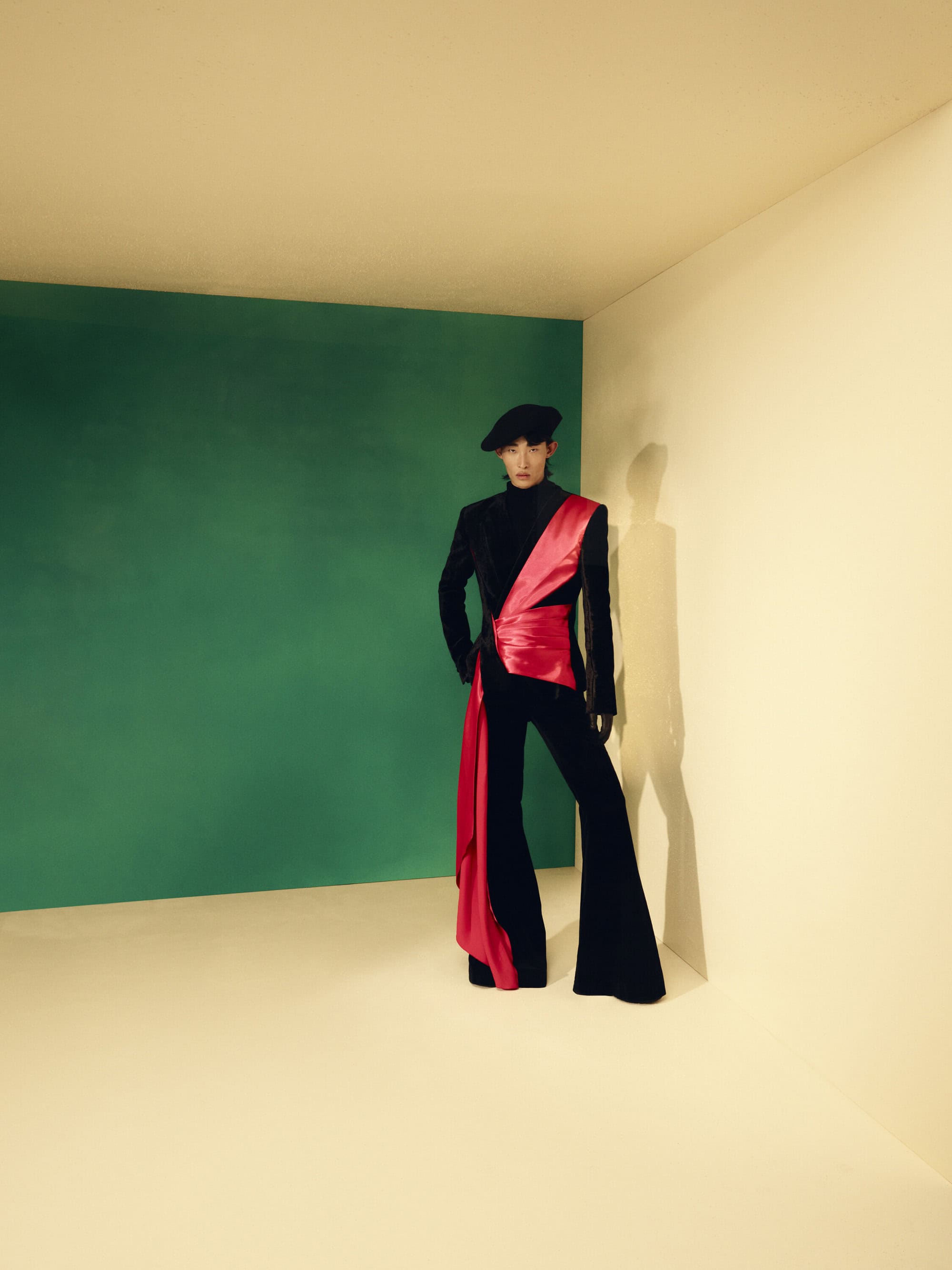 Stylish adult in a black suit with a striking red sash and matching beret strikes a pose in a minimalist studio setting with green and cream walls.