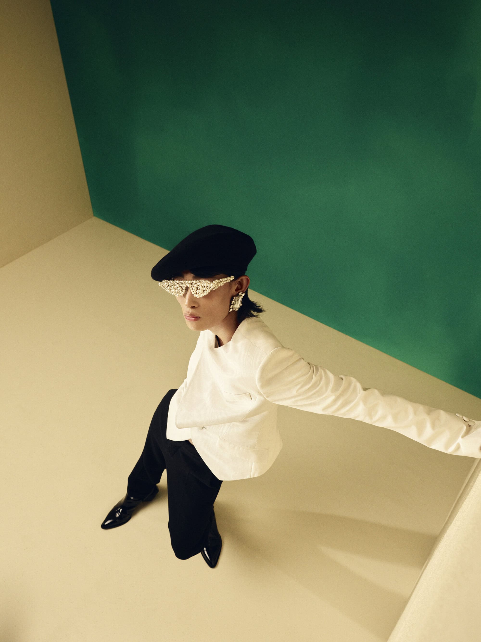 Stylish person in a white jacket, black pants and cap, and unique eyewear, posing in a studio with green and yellow wall.