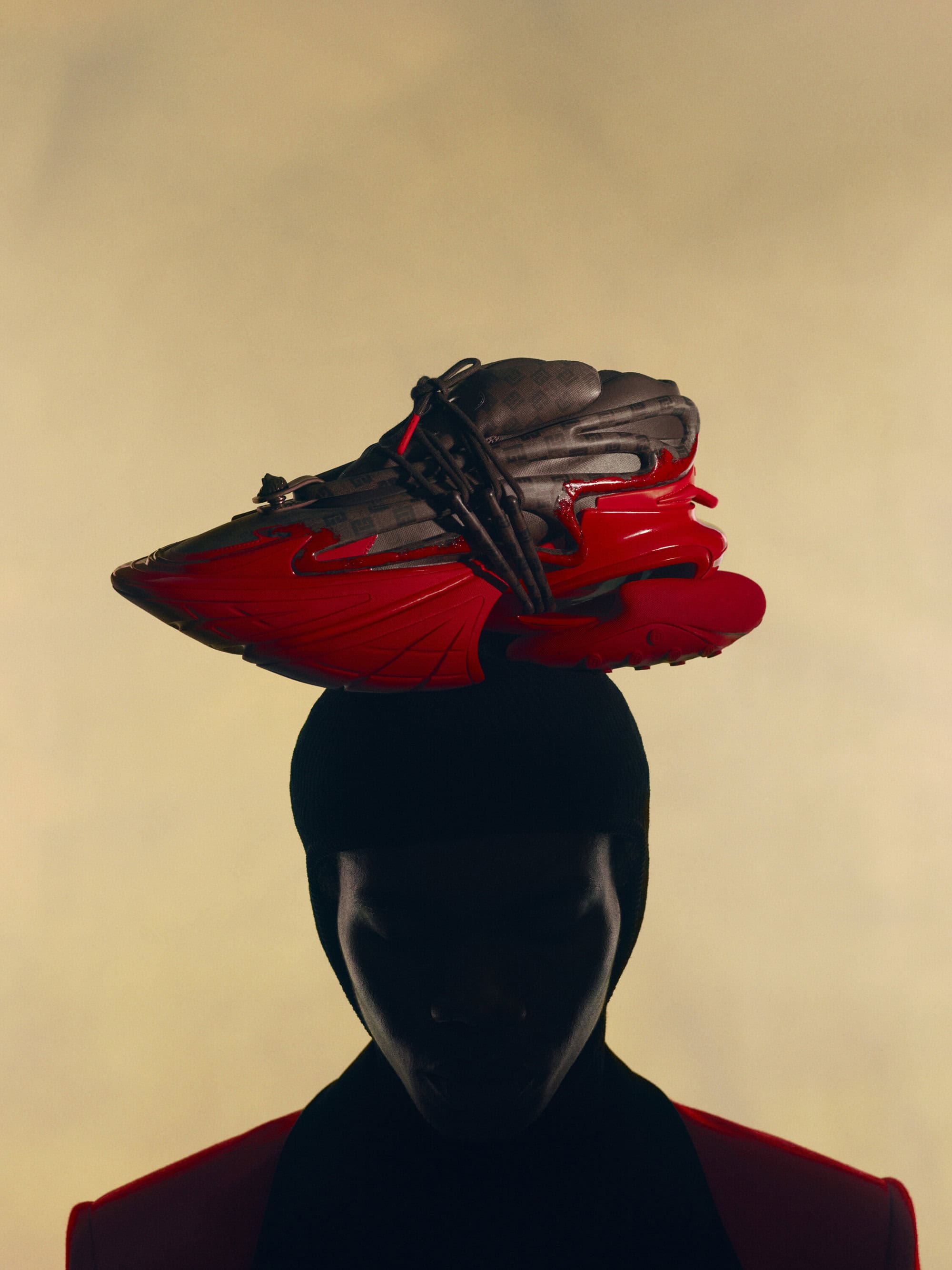 Person in silhouette poses with a black and red athletic shoe balanced on their head against a pale gradient background in studio lighting.