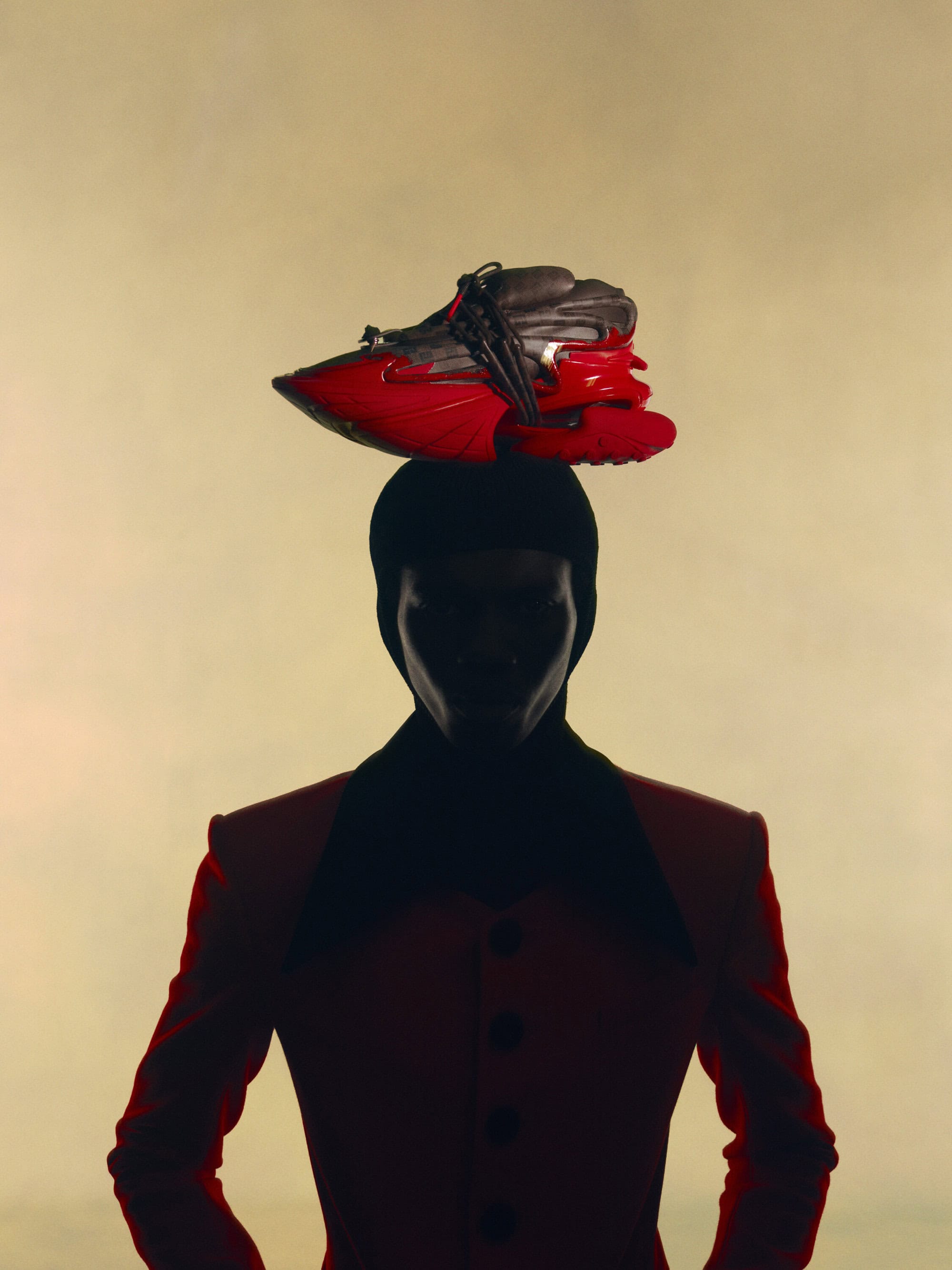 Silhouette of a person wearing a jacket, posing with a red and black sneaker balanced on their head in a brightly lit studio.