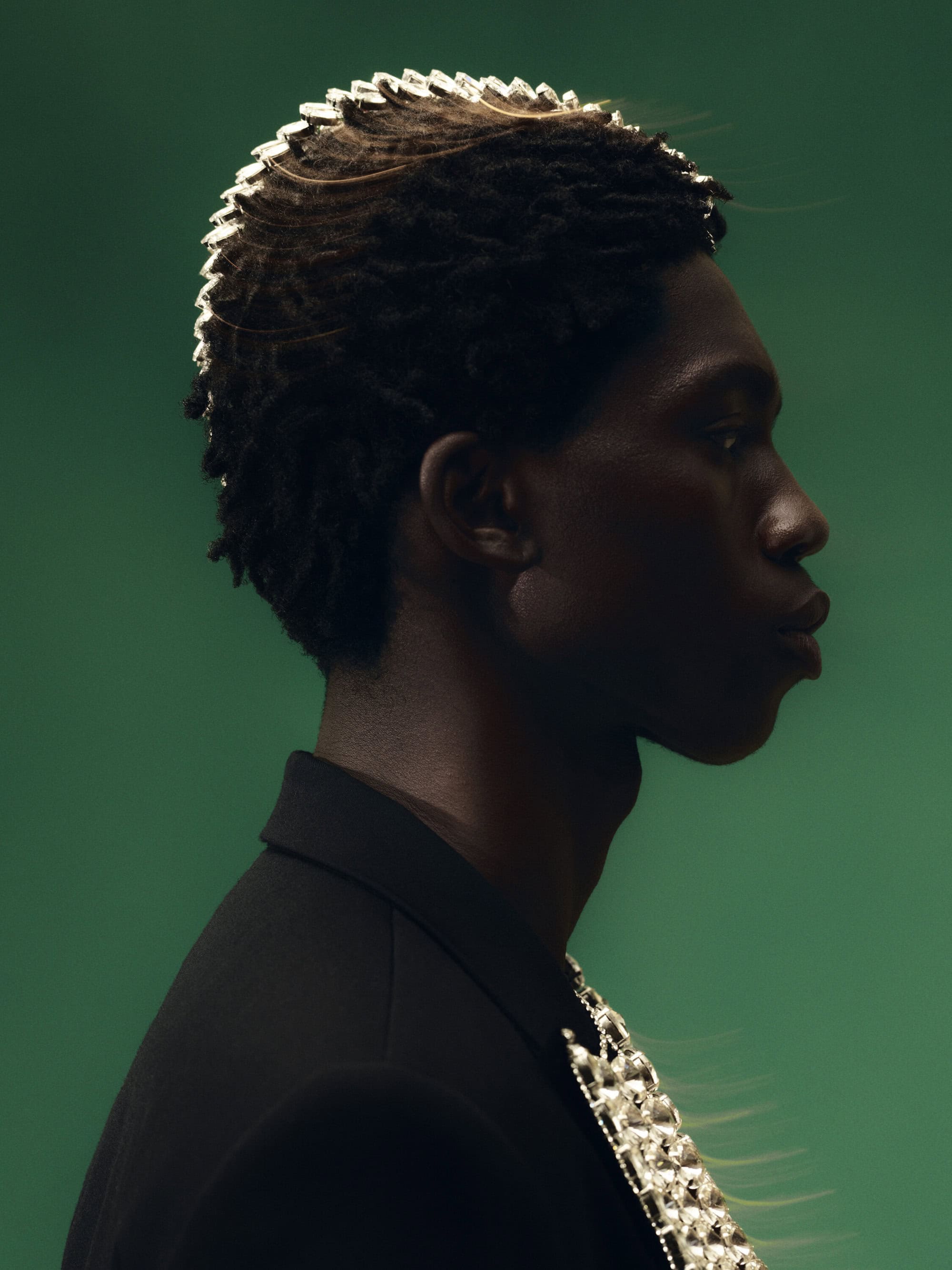Profile portrait of African-American man in black suit with metallic epaulettes against green backdrop in studio lighting.