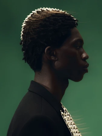 Profile portrait of African-American man in black suit with metallic epaulettes against green backdrop in studio lighting.