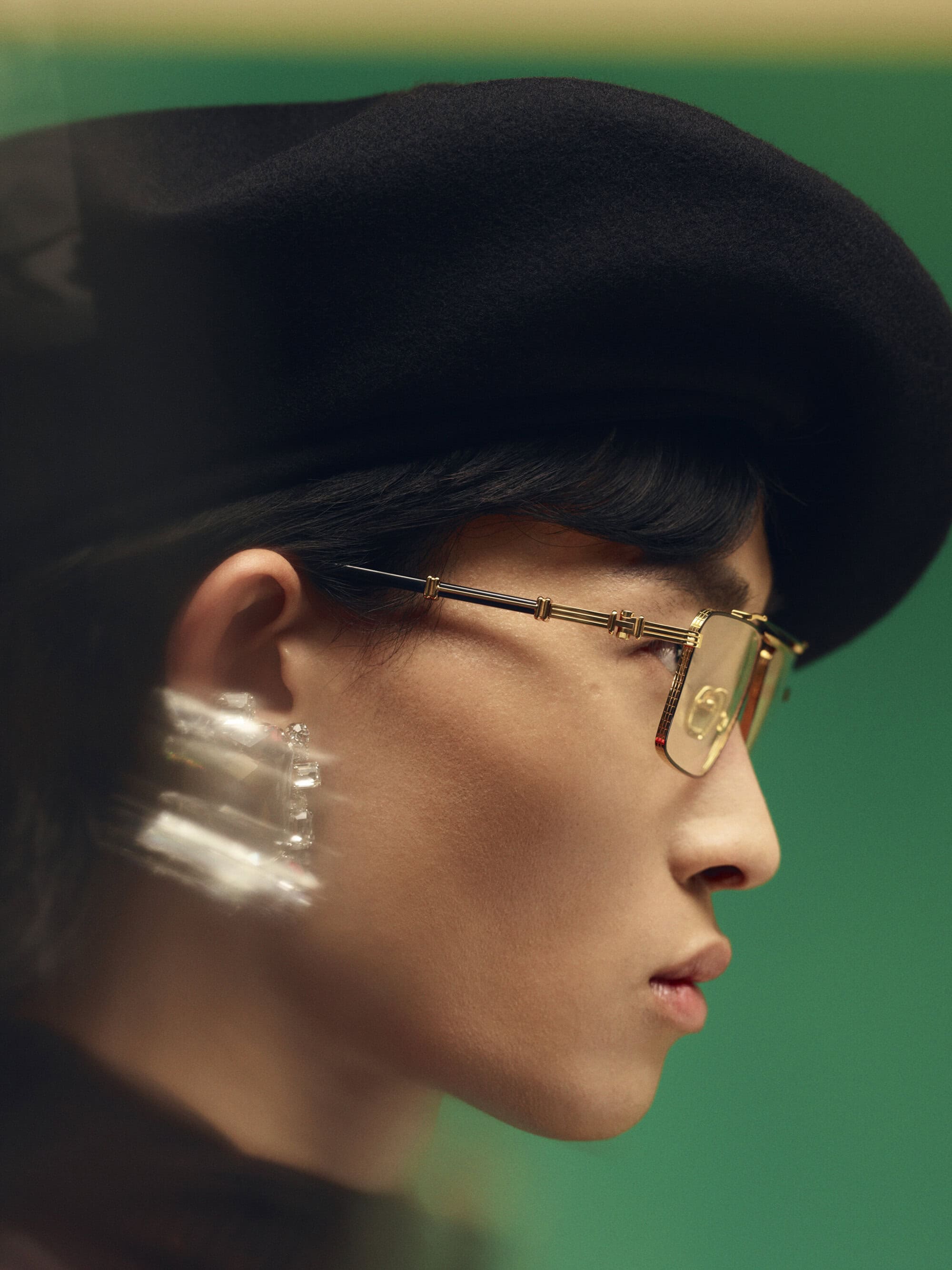 Close-up profile of an adult male wearing gold-framed eyeglasses and a black beret against a green background in a portrait-style shot.