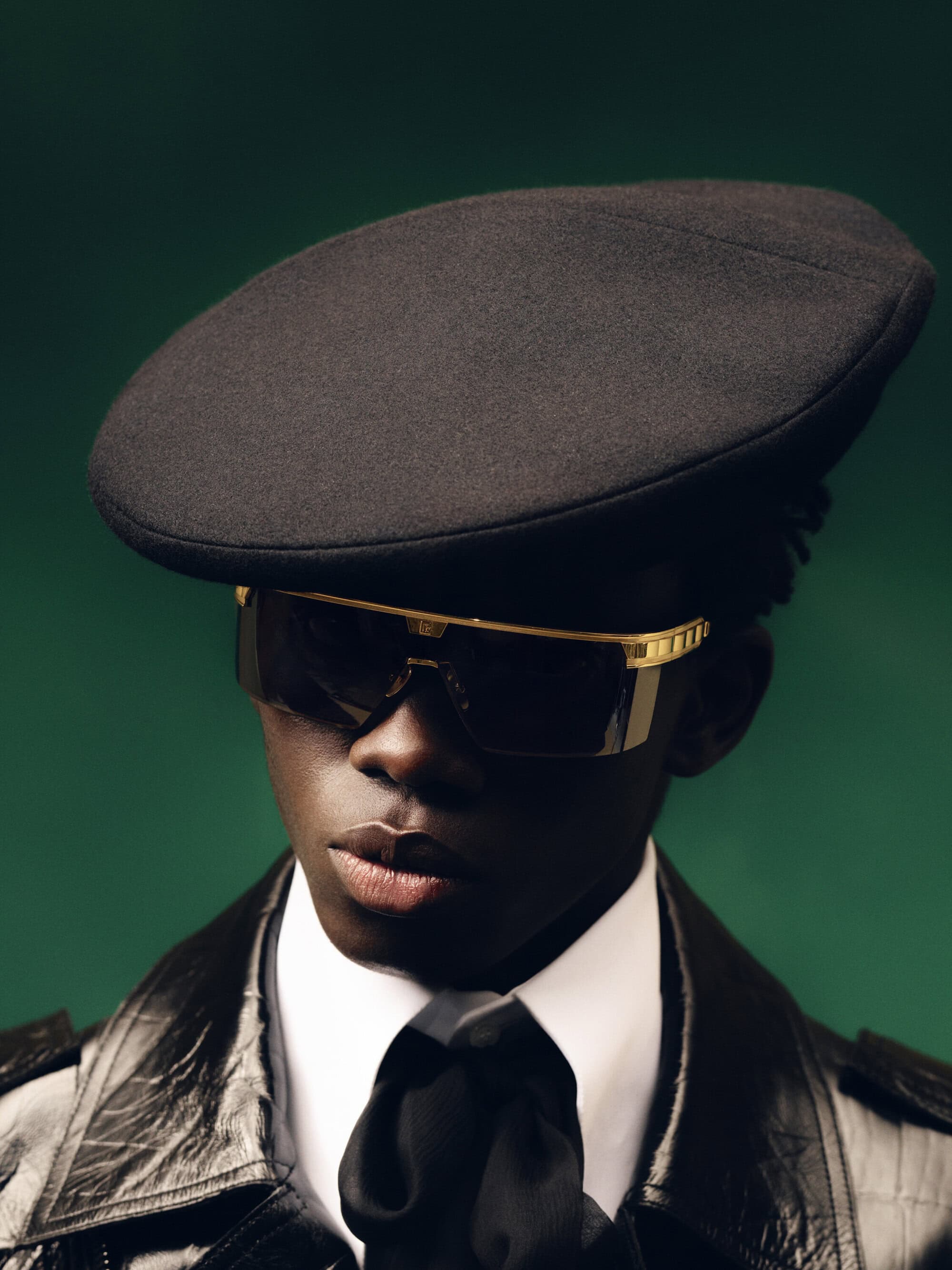 Dark-skinned model wearing a cap, sunglasses, and shiny black jacket against a solid dark green background in a close-up studio shot.