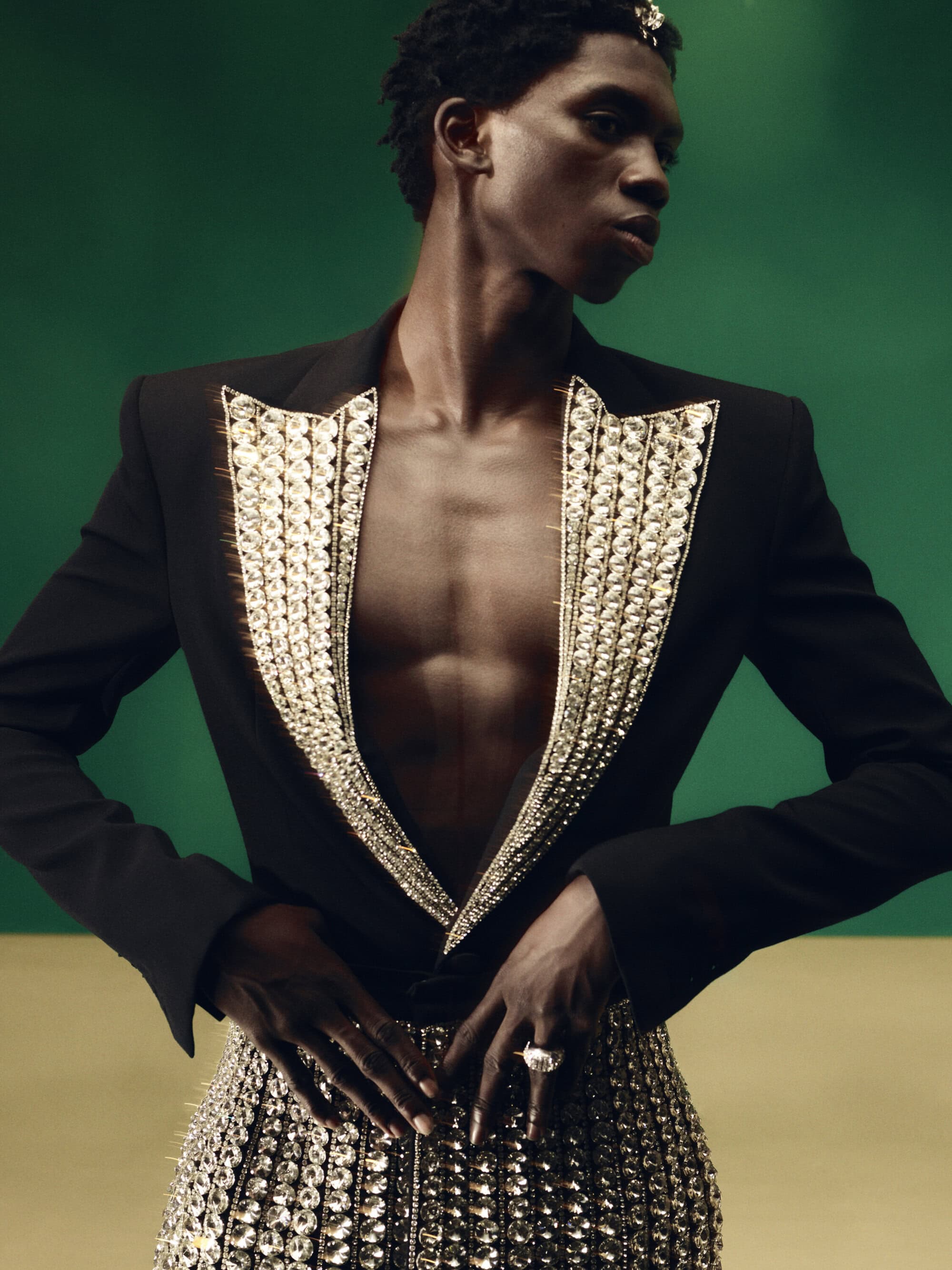 Fashionable Black male model in a black blazer, bejeweled bustier, and jewelry posing against a studio background with green and tan colors.