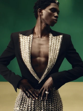 Fashionable Black male model in a black blazer, bejeweled bustier, and jewelry posing against a studio background with green and tan colors.