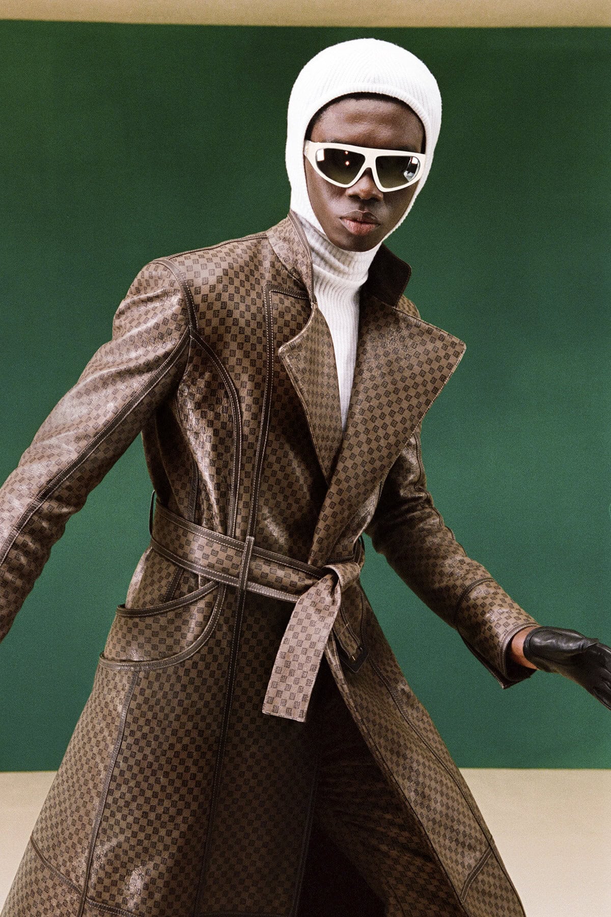 Fashion shot of a Black male model in a brown patterned coat, white turtleneck, white beanie, and sunglasses posing against a green backdrop.