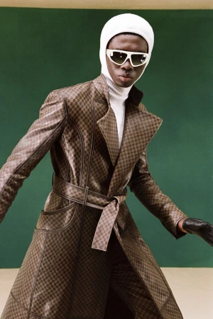 Fashion shot of a Black male model in a brown patterned coat, white turtleneck, white beanie, and sunglasses posing against a green backdrop.