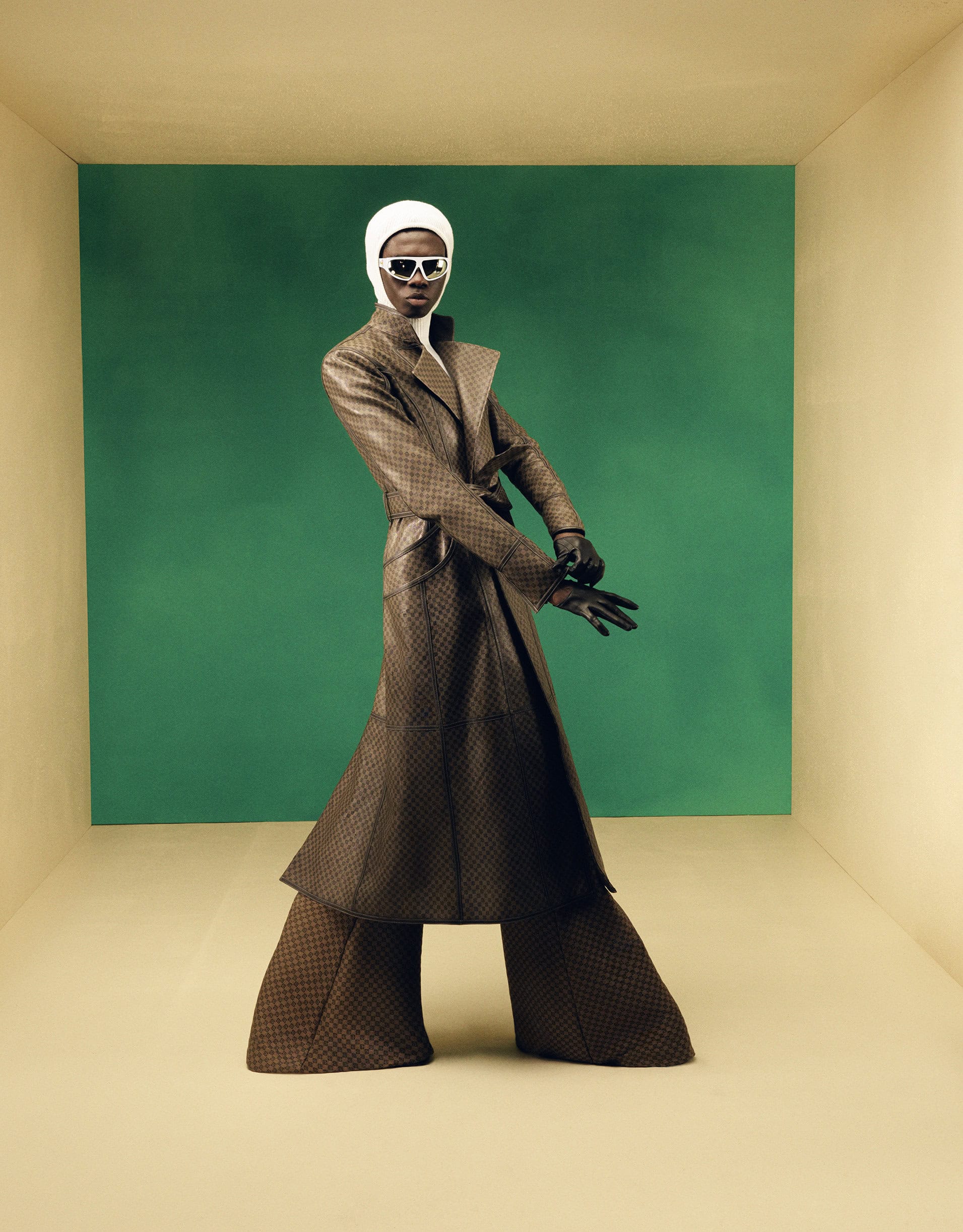Stylish adult posing in a tan room with a green panel, wearing a patterned brown coat, white head covering, black gloves, and sunglasses.