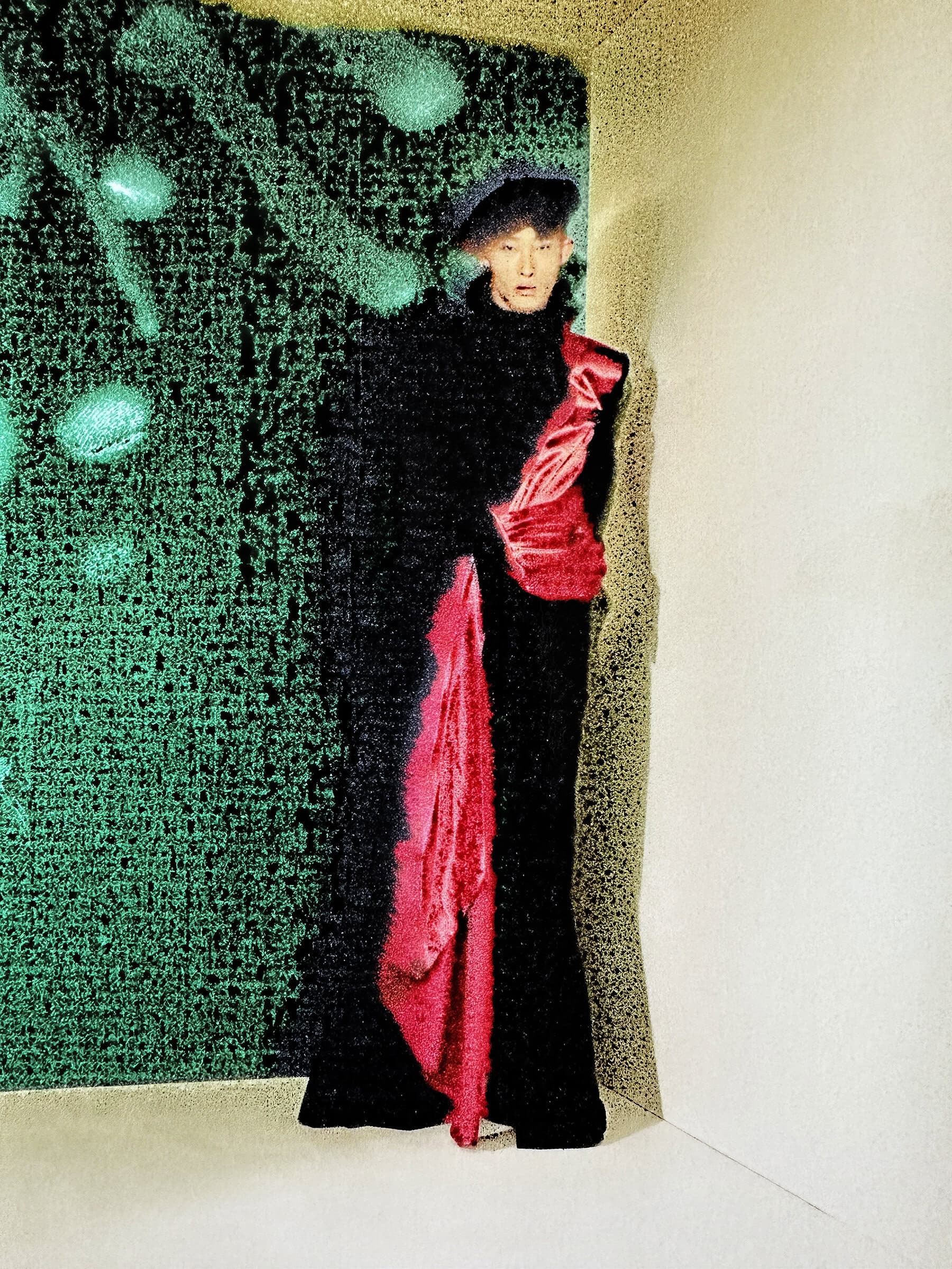 A person stands in a black outfit with a red sash against a green textured wall in an indoor studio setting with soft lighting.