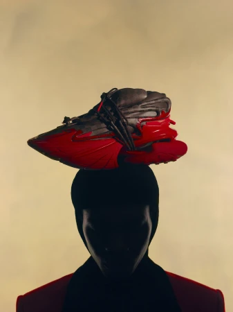 Silhouette of a person balancing an avant-garde red and black sculptural sneaker on their head against a warm neutral background.