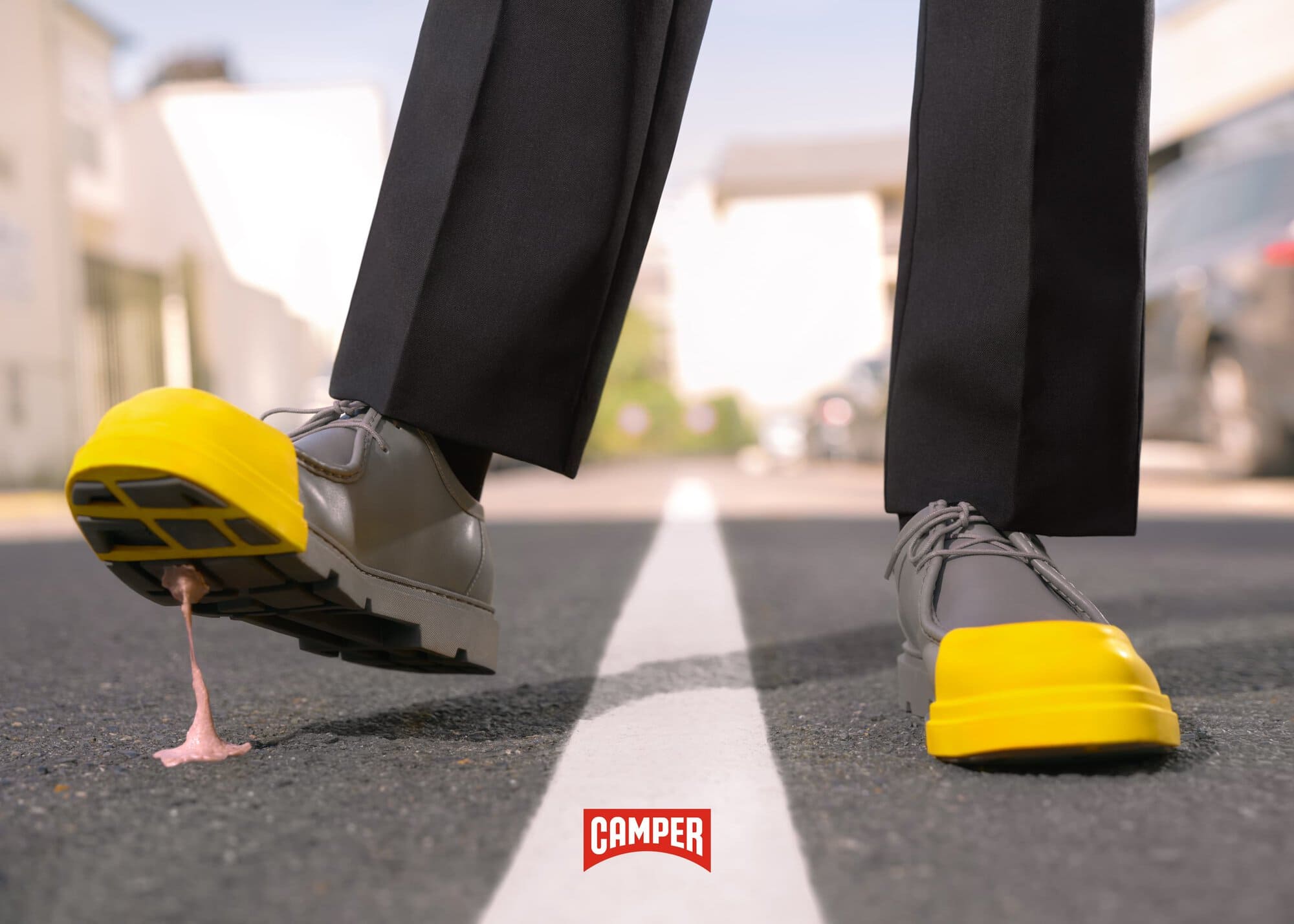 Footwear fashion shot: dress shoes with yellow treads stepping on pink bubble gum on pavement with shoe brand logo in foreground.