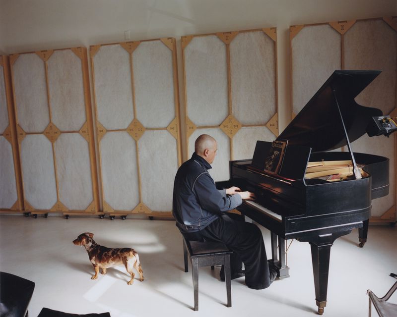 Bald adult man plays grand piano indoors as a dachshund dog looks on, with white soundproofing panels on the walls.