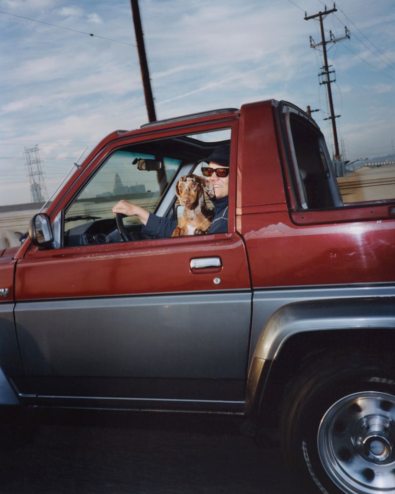 Man and dog ride in a red and gray SUV on a sunny day, with industrial and urban structures in the background, capturing a candid moment.