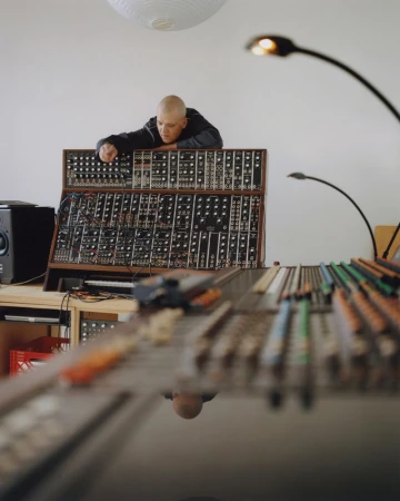 Bald man leans on an analog music synthesizer in a home studio with mixing consoles, studio monitors, and modern lighting fixtures.