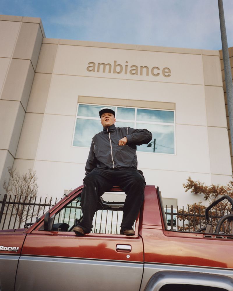 Young man sits on a red pickup truck under a building labeled ambiance in urban daylight, wearing dark clothing and hat.