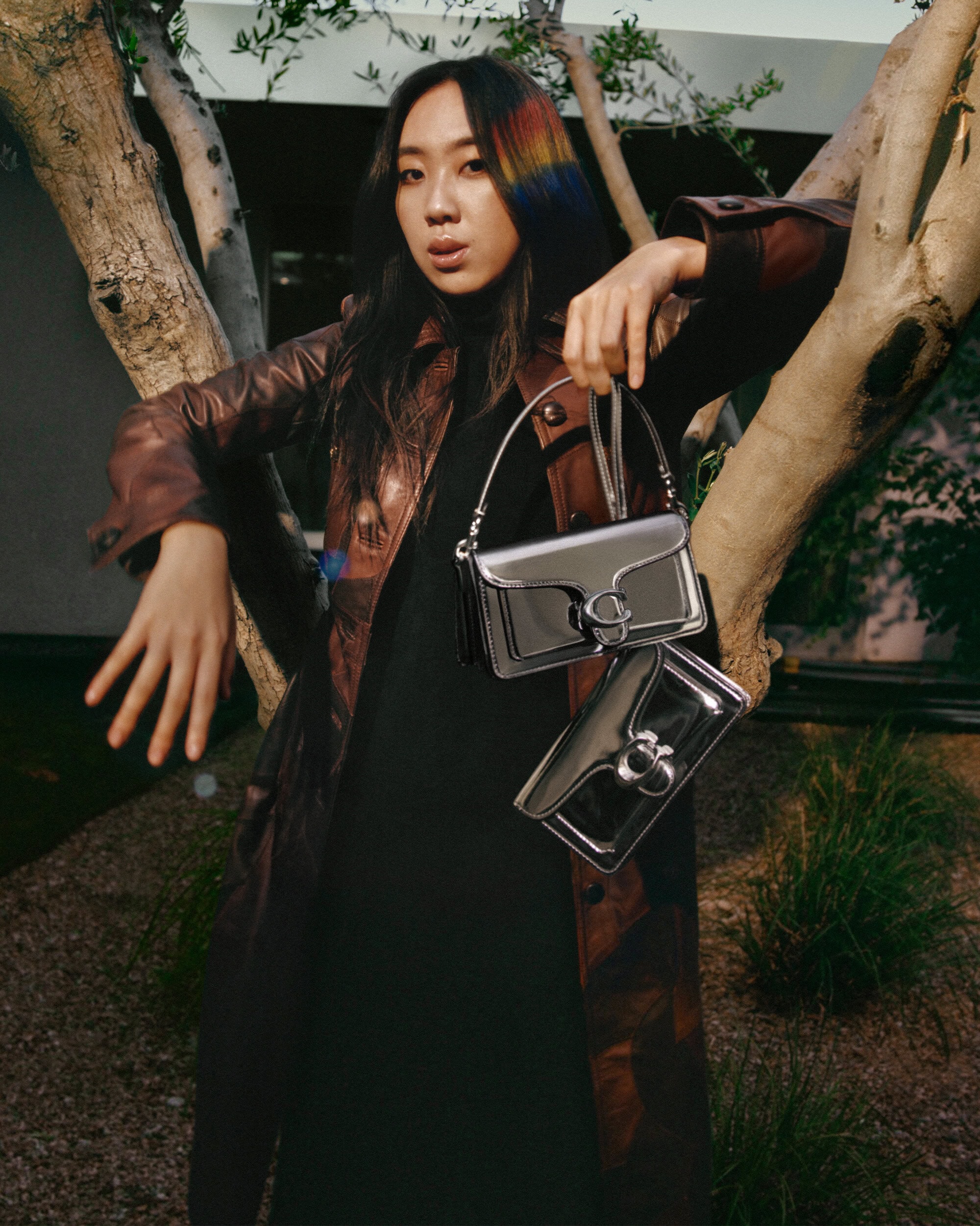 Asian woman with rainbow hair poses with two silver bags, wearing a dark dress and brown leather trench coat, in an outdoor setting.