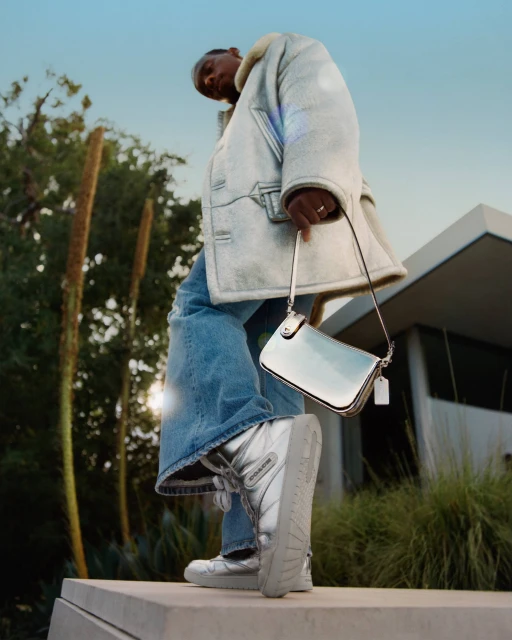 Fashion shot from a low angle of an adult wearing a silver jacket, jeans, and metallic boots holding a silver purse outdoors.