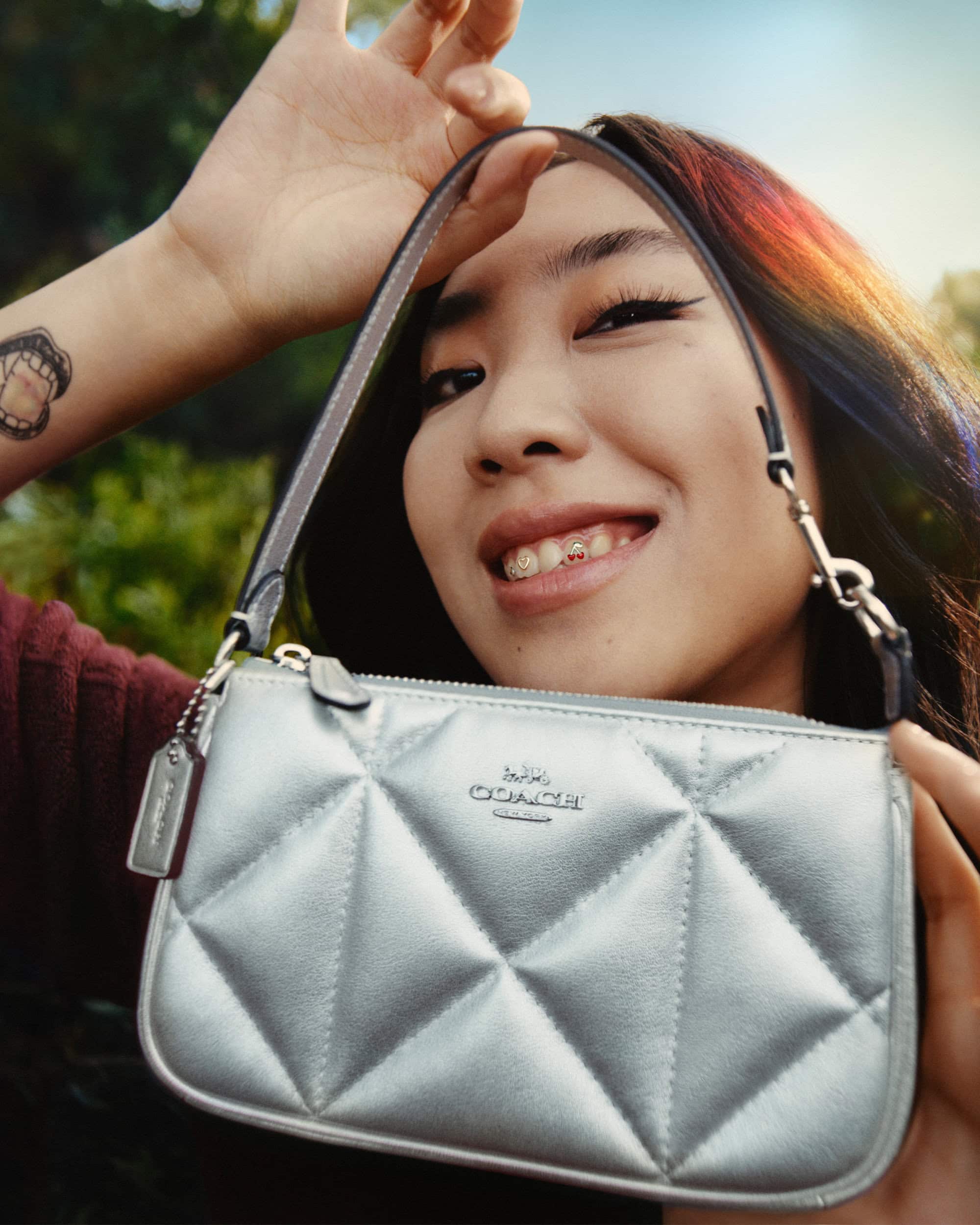 Smiling young woman displays a quilted silver Coach purse outdoors, showcasing the brand's logo and the purse's metallic sheen.