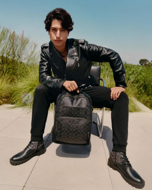 A stylish young man in a black leather jacket poses with a Coach backpack outdoors, complemented by black trousers and leather boots.