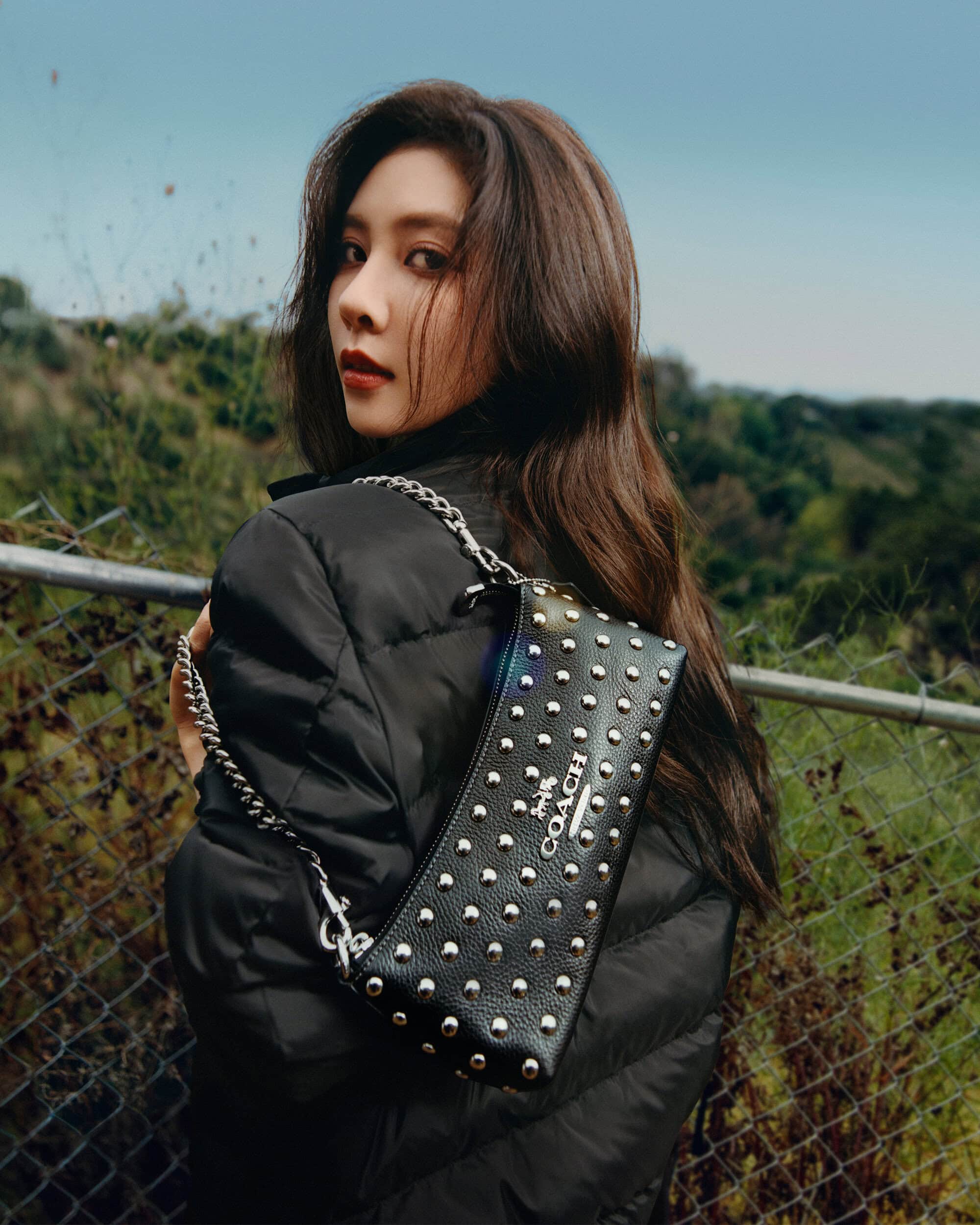 Adult woman with long brown hair poses outdoors with a studded Coach bag; she wears a black puffer jacket in a medium shot.