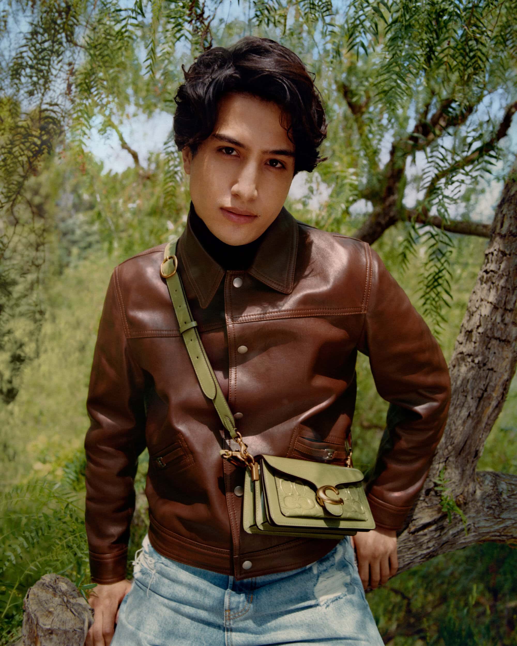 A stylish young adult poses outdoors in a leather jacket, light jeans, and a crossbody bag, standing against a backdrop of leafy trees.