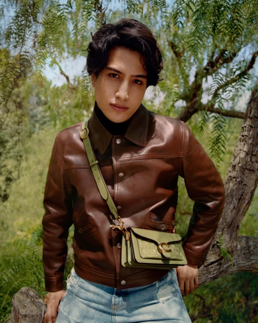 A stylish young adult poses outdoors in a leather jacket, light jeans, and a crossbody bag, standing against a backdrop of leafy trees.