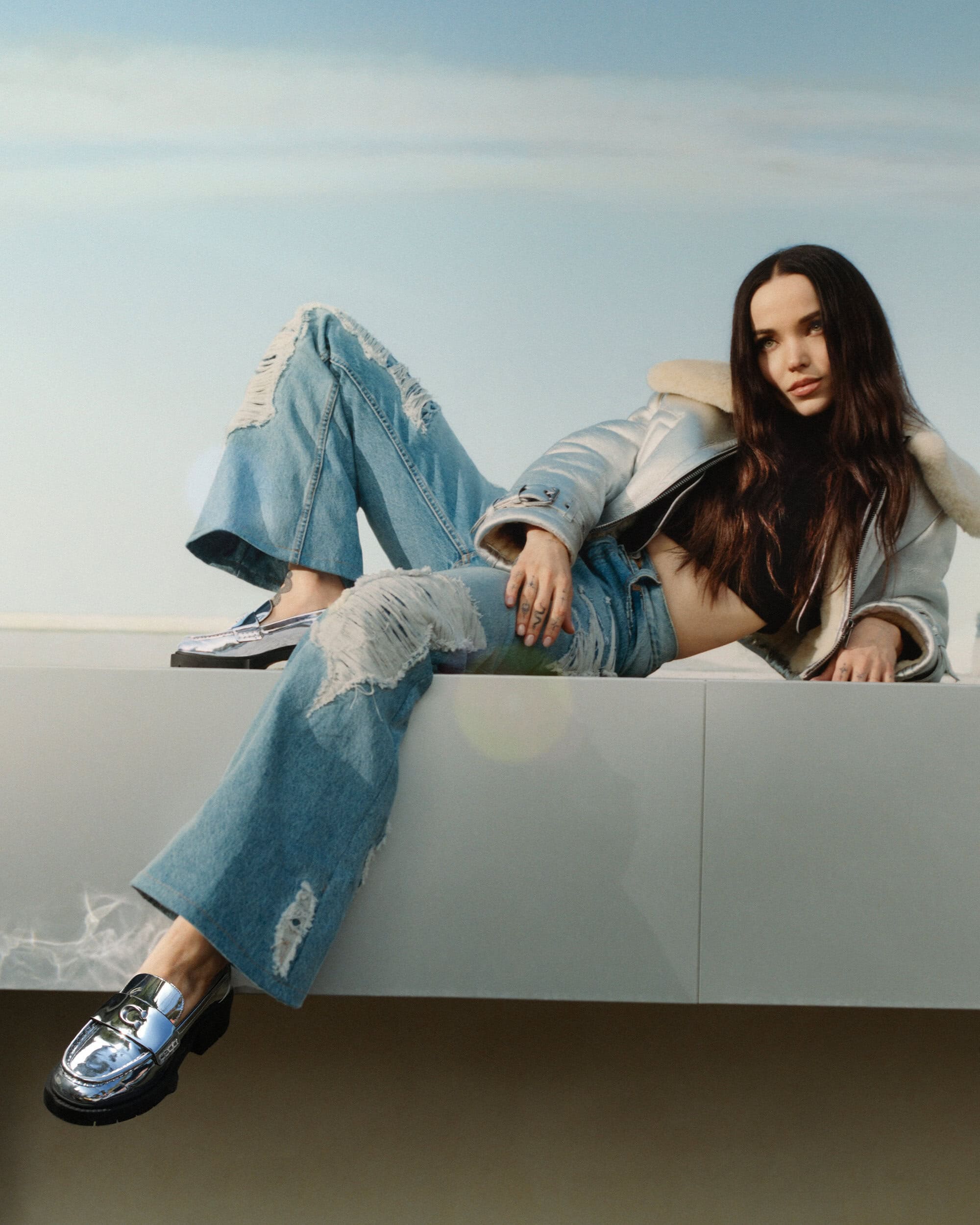 A young woman poses outdoors, wearing a light blue denim jacket with fur collar, ripped jeans, and silver loafers against a light blue sky.
