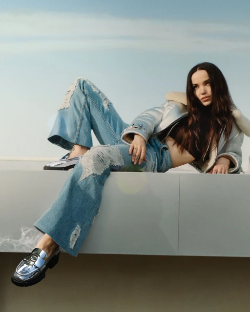 A young woman poses outdoors, wearing a light blue denim jacket with fur collar, ripped jeans, and silver loafers against a light blue sky.