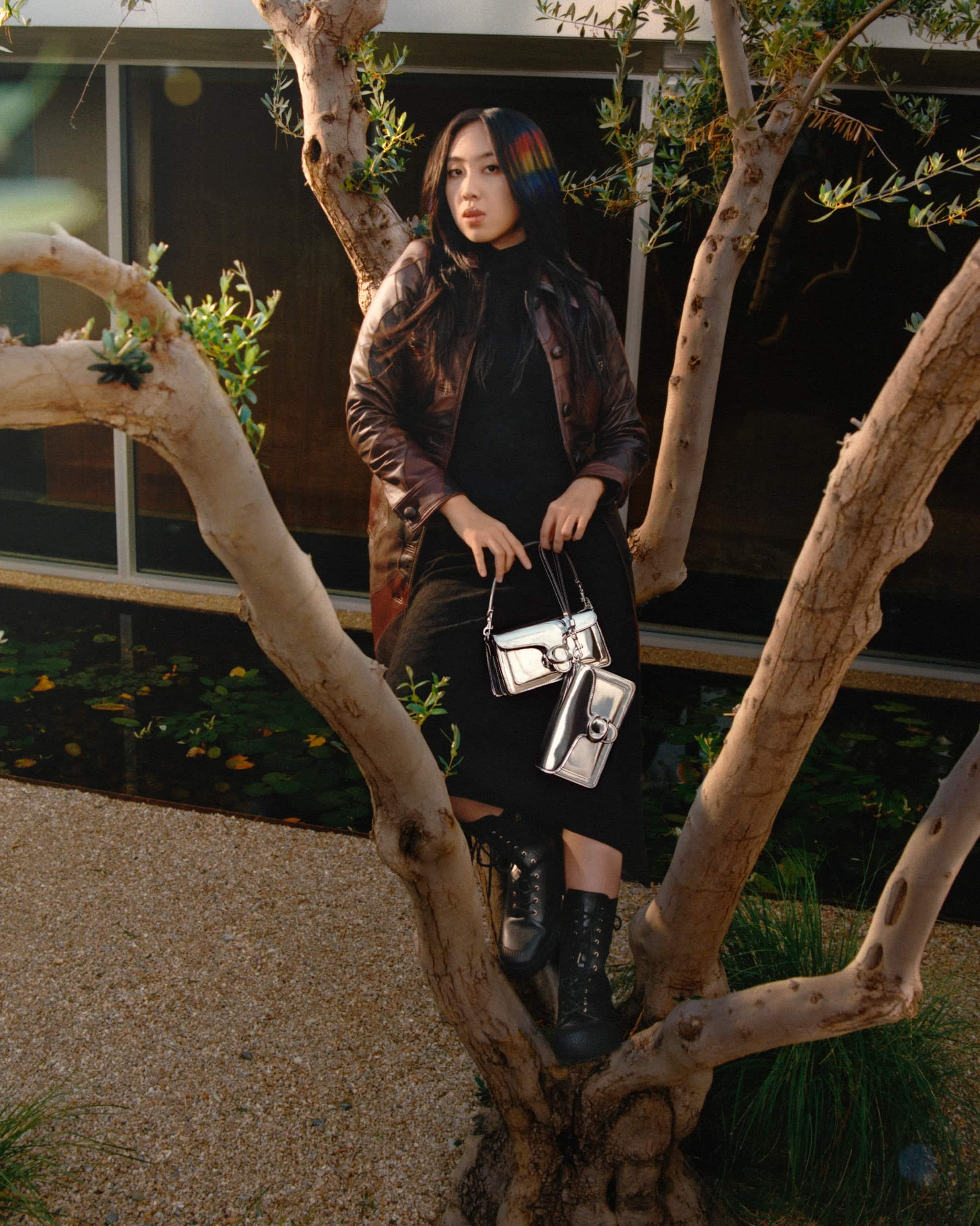 A stylish Asian woman poses on a tree branch wearing a brown jacket, black dress, and boots while holding silver handbags.