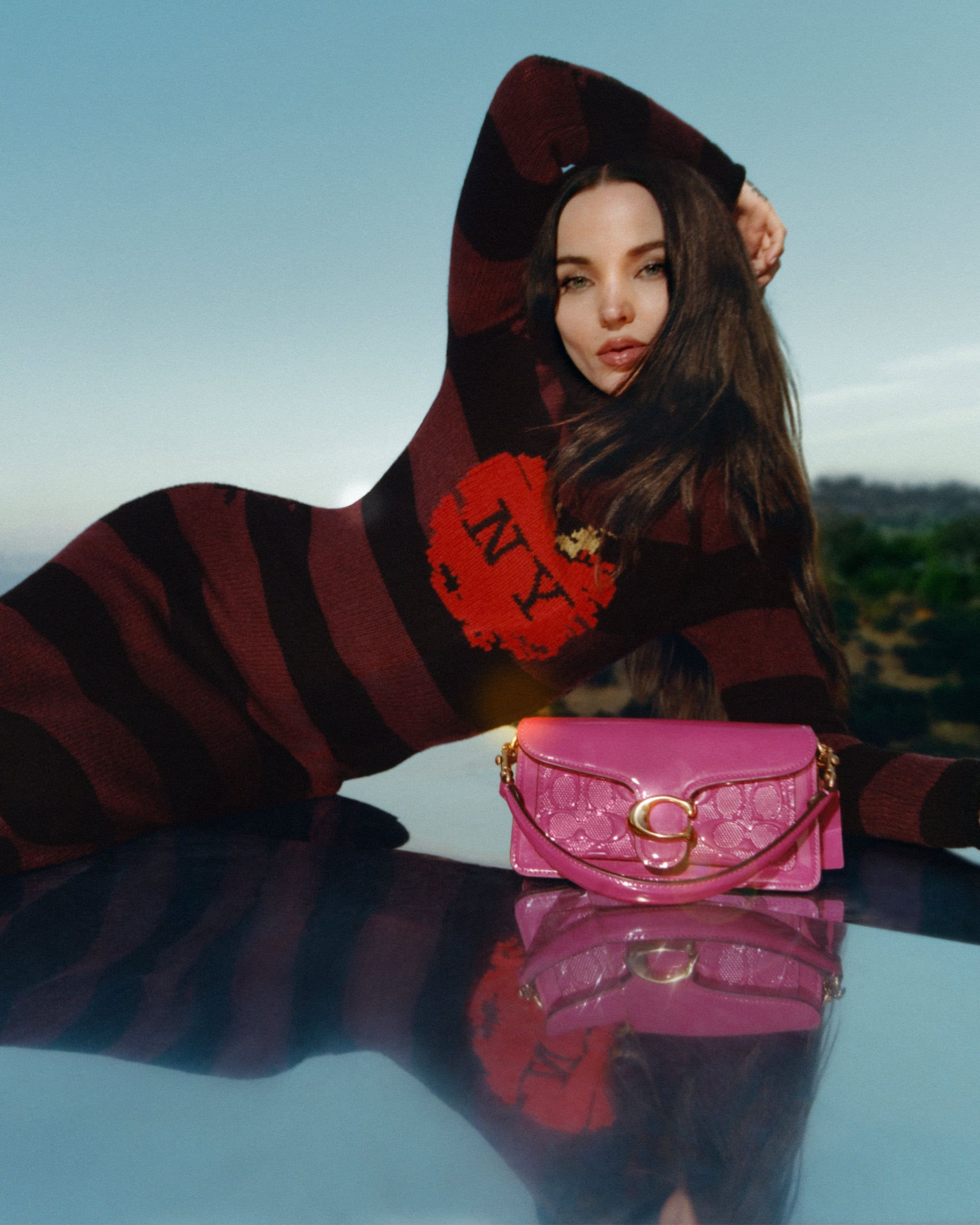 Woman poses with a Coach bag outdoors, wearing a maroon striped dress with 'NY' on it, reflecting on a shiny surface against blue skies.