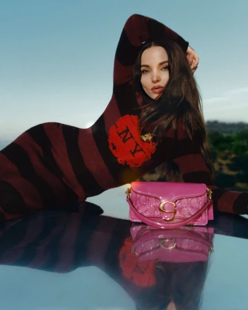 Woman poses with a Coach bag outdoors, wearing a maroon striped dress with 'NY' on it, reflecting on a shiny surface against blue skies.