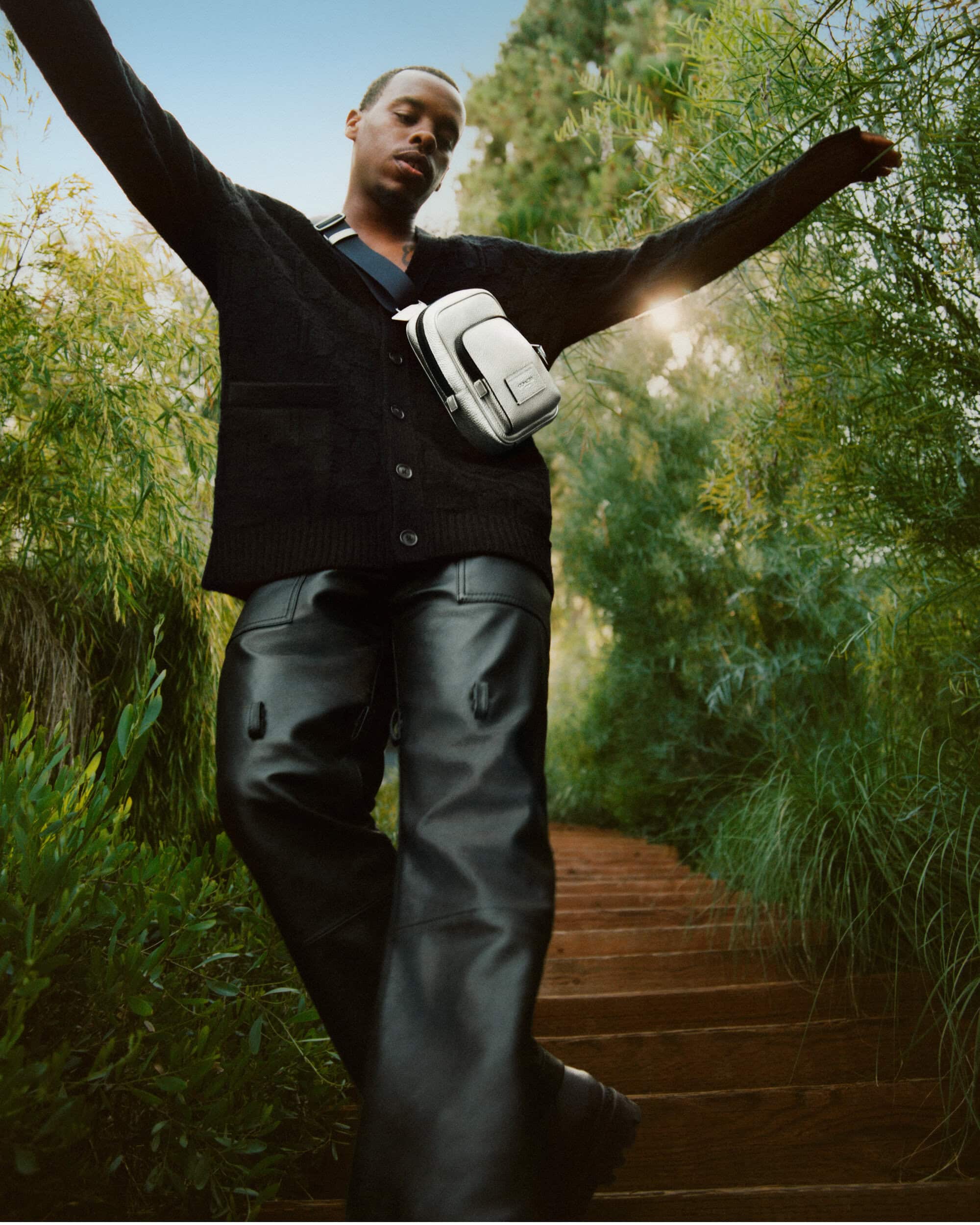 An adult male model wears a dark cardigan, black leather pants and a white crossbody bag while standing on outdoor steps in a green setting.