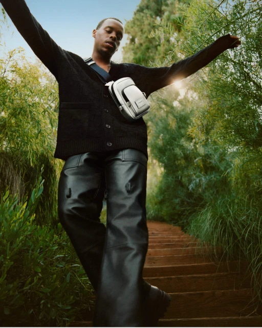 An adult male model wears a dark cardigan, black leather pants and a white crossbody bag while standing on outdoor steps in a green setting.