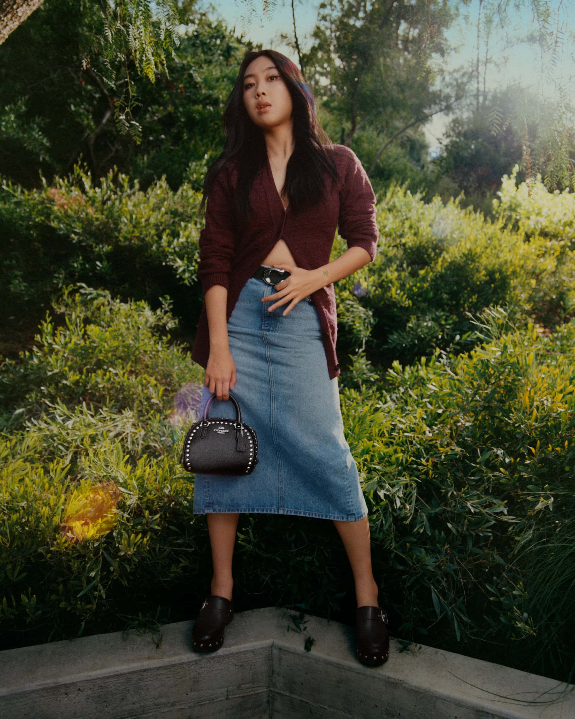 Adult woman in a v-neck cardigan, denim skirt, and clogs, holding a black dome satchel with studs, poses in a sunny garden setting.